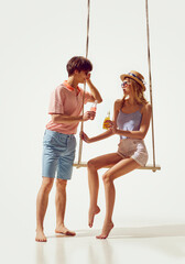 Wall Mural - Young man cheerfully talking with beautiful young woman in swimsuit, people drinking lemonade against white background. Concept of summer vacation, travelling, retro style, fashion