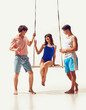 © master1305 - Beautiful young woman in blue swimsuit sitting on swing and cheerfully talking to two handsome young men against white background. Concept of summer vacation, travelling, retro style, fashion
