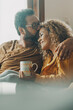 © simona - Couple of young adult man and woman enjoy time together sitting on the sofa in indoor leisure activity.. Real life people at home. Male and female relax on the couch reading a book and drinking a tea