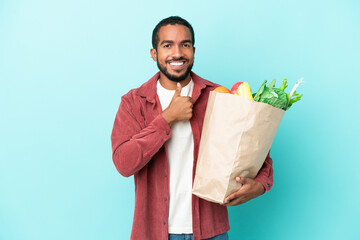 Sticker - Young latin man holding a grocery shopping bag isolated on blue background giving a thumbs up gesture