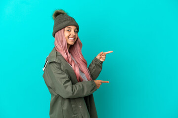 Wall Mural - Young woman with pink hair wearing a rainproof coat isolated on blue background pointing finger to the side and presenting a product