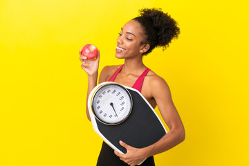 Wall Mural - Young African American woman isolated on yellow background with weighing machine and with an apple