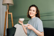 A cheerful pretty young woman with a cup of hot drink sitting on a couch enjoying a vacation. Couch Comfort. Cheerful Woman Savoring Hot Beverage on Holiday.
