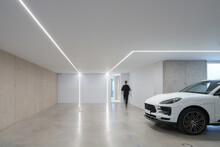 Unrecognizable person walking in contemporary garage with integrated LED lighting, polished concrete floor, and a parked luxury car