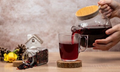 Wall Mural - A woman's hand pours tea from a glass transparent jug into a cup. Hot healthy drink, fruit tea in a transparent glass.