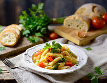 Pasta With Vegetables On A White Plate Stands On A Wooden Textured Plank And Textiles, In The Background Is Soddy Rustic Bread And Cilantro