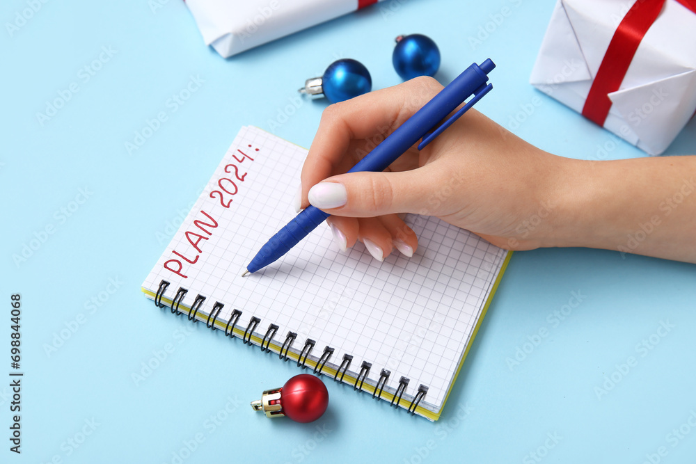 Woman writing PLAN 2024 in notebook and Christmas decor on blue background, closeup