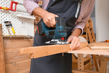 Mature carpenter sawing wooden board with jigsaw at table in workshop, closeup