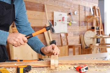 Canvas Print - Mature carpenter hammering nail into wooden plank at table in shop, closeup