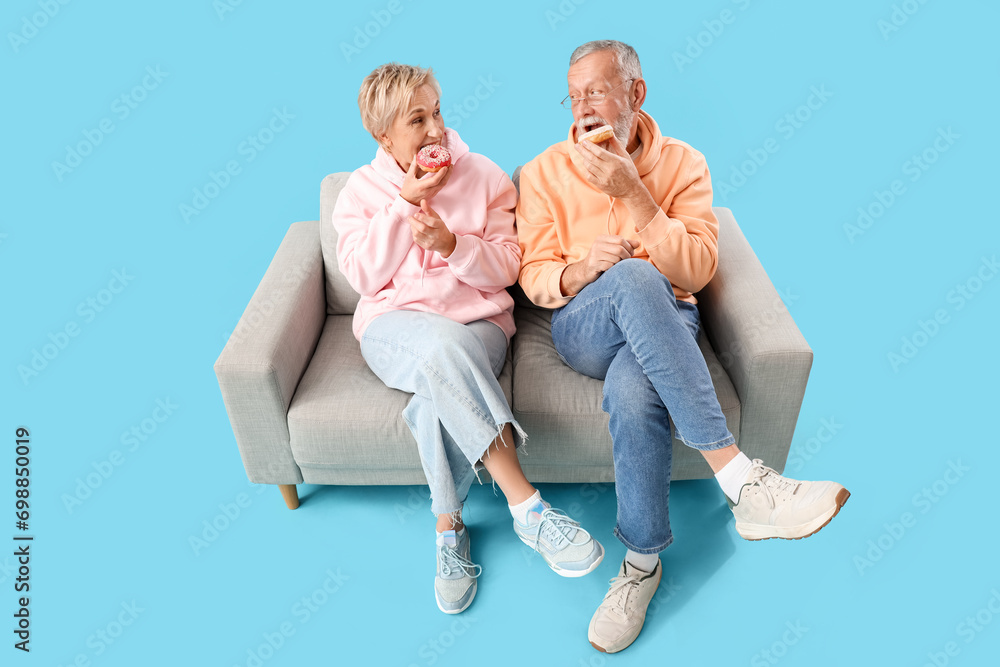 Mature couple eating tasty donuts on sofa against blue background