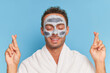 © South House Studio - Young man with towel on his shoulders and clay mask on his face holding hands up with crossed fingers, spa treatments concept, copy space