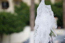 Water Fountain Geyser Caught Midair, White Water Foam Frozen In Time, Foliage In Unfocused Background