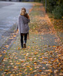 Woman walks along a sidewalk covered with autumn leaves, view from behind while walking away.