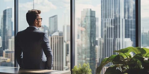 Wall Mural - Back View of the Thoughtful Businessman wearing a Suit Standing in His Office, Hands in Pockets and Contemplating Next Big Business Deal, Looking out of the Window. Big City Business District View
