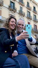 Wall Mural - Cheerful mid adult couple having fun together taking a selfie in the street. Romantic senior couple smiling taking picture together in european city.