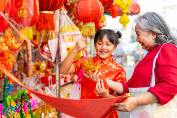 Happy Asian family grandmother and grandchild girl in red dress choosing and buying home decorative ornaments for celebration Chinese Lunar New Year festival together at Chinatown street market.