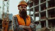 Cute bearded construction worker with safety helmet on head in vest standing with arms crossed at construction site and looking at camera
