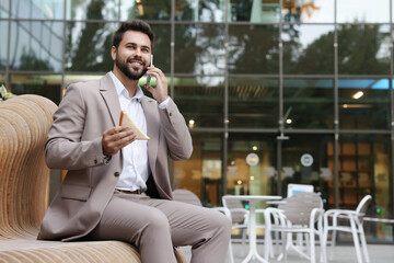 Wall Mural - Lunch time. Young businessman with sandwich talking on smartphone on bench outdoors