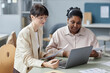 © Seventyfour - Medium shot of two smiling diverse female coworkers holding papers looking at laptop while sitting at desk in office