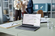 © Seventyfour - Medium shot with focus on opened laptop displaying business report infographic, water glasses and clipboards on office table, unrecognizable company employees standing in blurred background