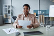 © Seventyfour - Portrait of adult African American businesswoman wearing pastel suit jacket holding paper while sitting at office desk and looking at camera