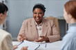 © Seventyfour - Medium shot of laughing African American female candidate wearing pastel suit gesturing and telling about herself during job interview with unrecognizable HR agents in office