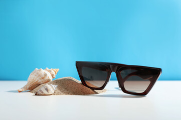 Poster - Stylish sunglasses, seashells and sand on white table against light blue background. Space for text