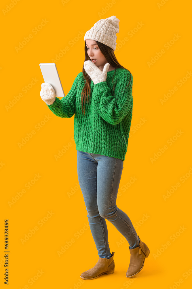 Shocked young woman in warm hat using tablet computer on yellow background