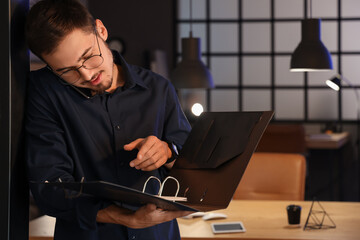 Wall Mural - Handsome young man with folder talking by mobile phone in office at night