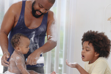 Happy African family, toddle baby infant sits on towel after takes a bath in bathroom, father and son brother hold dusting powder bottle and apply talcum powder on body of little kid daughter child.