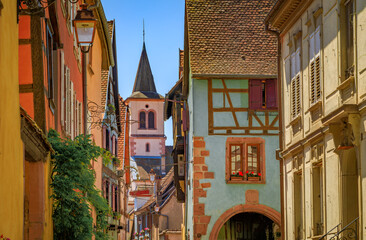 Canvas Print - View onto a church tower and ornate traditional half timbered houses with blooming flowers in a village on the Alsatian Wine Route, Riquewihr, France