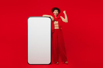 Wall Mural - Full body young woman wears beige sweater hat scarf big huge blank screen mobile cell phone smartphone with area do winner gesture isolated on plain red background studio portrait. Lifestyle concept.