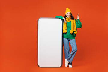 Wall Mural - Full body young happy woman she wear green knitted sweater yellow hat scarf big huge blank screen mobile cell phone smartphone with area point index finger up isolated on plain orange red background.