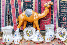 A Fake Dromedary And White Ceramic Pottery Jugs With Colorful Berber Motifs And Two Plates With A City And A Mosque Minaret And A Tuareg Man Pouring Tea. Berber Carpets In Foreground And Background.