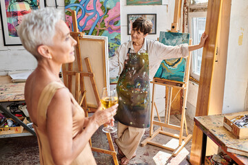 Wall Mural - happy woman artist looking at elegant middle aged model posing with wine glass in art workshop