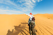 A young women  rides a camel through the dunes in the Sahara Desert. View of the woman from behind, in the background, small silhouettes of other tourists. Merzouga, Morocco