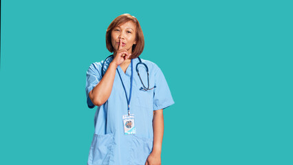 Wall Mural - Portrait of stern serious asian nurse shushing at camera, bothered by excessive noise. Strict healthcare manager holding quiet gesture finger to lips, isolated over blue studio background