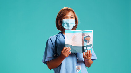 Wall Mural - Experienced nurse presenting medical leaflet containing list of clinic professional services. Hospital representative advertising healthcare brochure, isolated over blue studio background