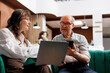 © DC Studio - Caucasian elderly couple using their laptop and mobile phone in hotel lobby, waiting for check-in procedure. Female tourist with laptop making online reservation with male traveler holding smartphone.
