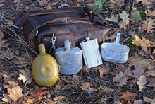 A Row Of Three Old Flasks And An Aluminum Pot Stand On The Ground Next To A Brown Backpack On The Street