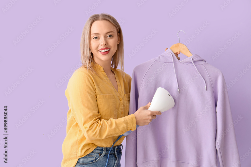 Pretty young woman steaming clothes with modern steamer on lilac background