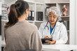 © M+Isolation+Photo - An elderly Asian doctor is talking to a younger Asian woman across a desk in a medical office, monthly health check appointment.