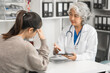 An elderly Asian doctor is talking to a younger Asian woman across a desk in a medical office, monthly health check appointment.