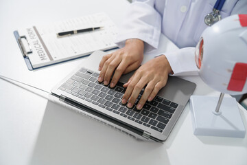 A doctor's hands are seen typing on a laptop keyboard with a stethoscope around the neck on white desk.