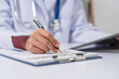 A doctor's hand is writing on a medical record form on a clipboard, with a stethoscope and tablet in the background.