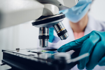 A scientist examines samples with a microscope in a laboratory with test tubes filled with colorful liquids in the background.