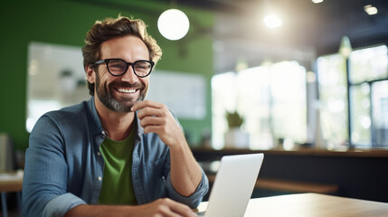 Poster - Cheerful man with a beard and glasses working on a laptop at a wooden desk