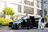 Fototapeta Przestrzenne - Female chauffeur waits a business people to let them in a minivan taxi, keeping door open. Concept of business trips and transportation service