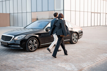 Wall Mural - Businessman and businesswoman walk together to car on parking lot near modern building, going to drive by luxury car