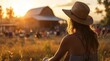 a woman outdoors, sitting in a straw hat watching an old barn at sunset